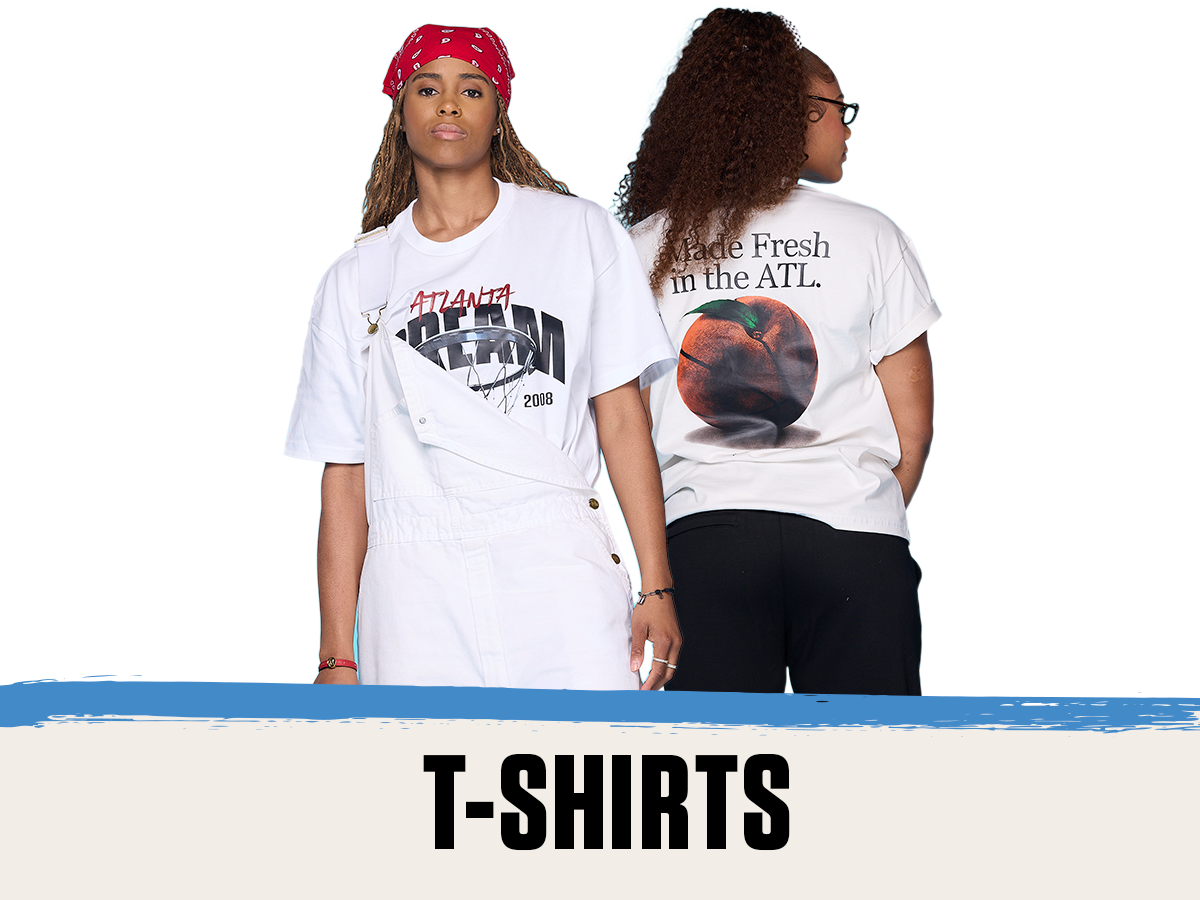 Two players model Atlanta Dream graphic t-shirts. The woman on the left wears a white tee with a bold "ATLANTA DREAM" basketball design and red bandana, styled under white overalls. The woman on the right shows the back of her shirt, which features a large peach basketball graphic with the phrase "Made Fresh in the ATL." The image promotes the T-SHIRTS section with a bold title at the bottom.